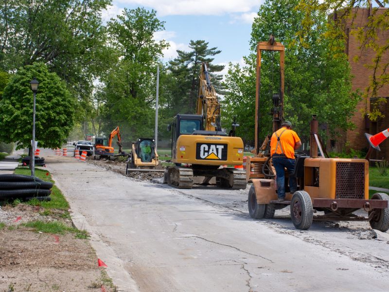 Construction workers work on a road project, they are sitting on equipment and construction barrels can be seen